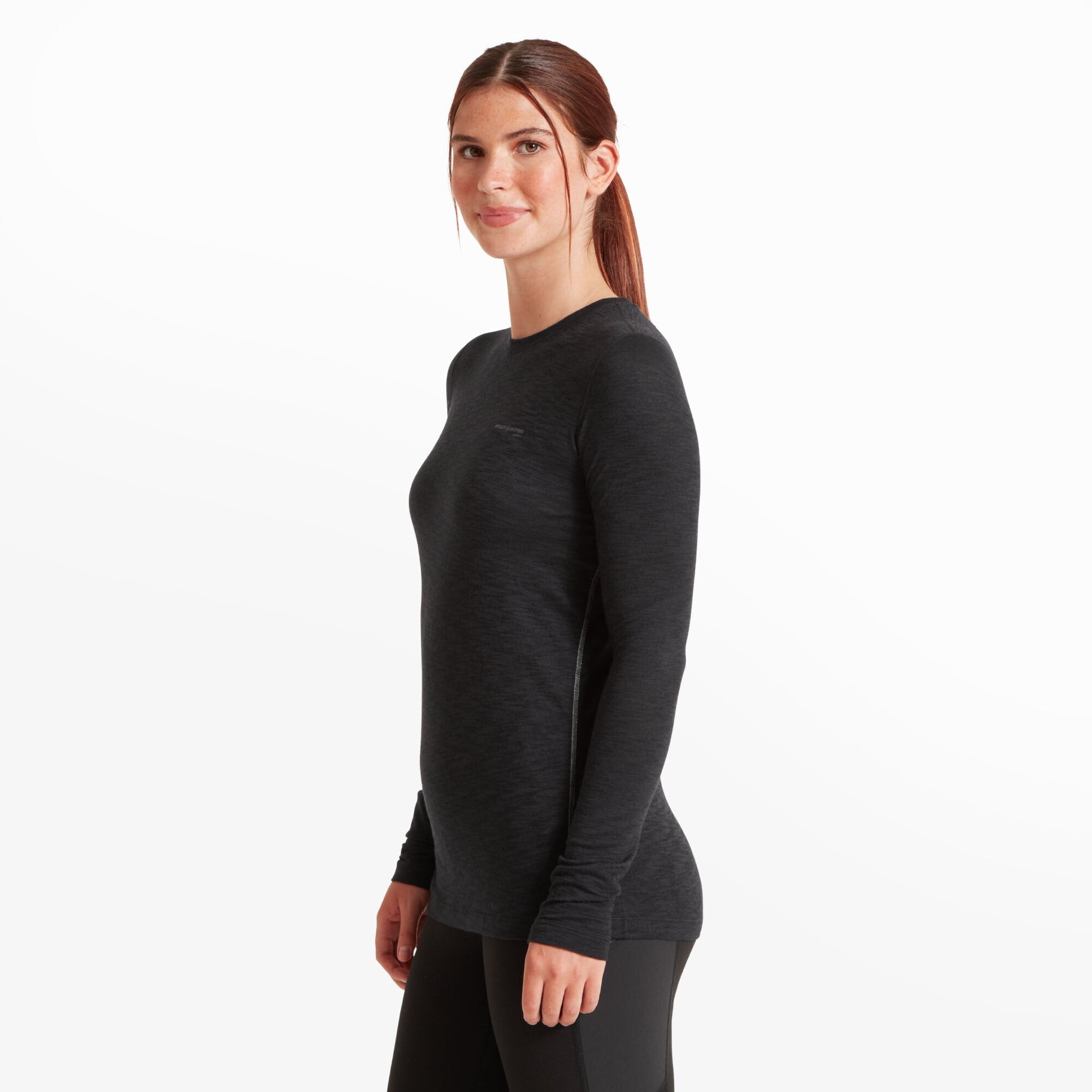 Side profile view of the same woman wearing the Artilect Performance Womens Sprint Long Sleeve Tee Black. The shirt’s slim fit and ergonomic seam placement are visible, showcasing the contoured silhouette and tailored fit through the torso and sleeves. Her posture is upright, with a gentle smile on her face.