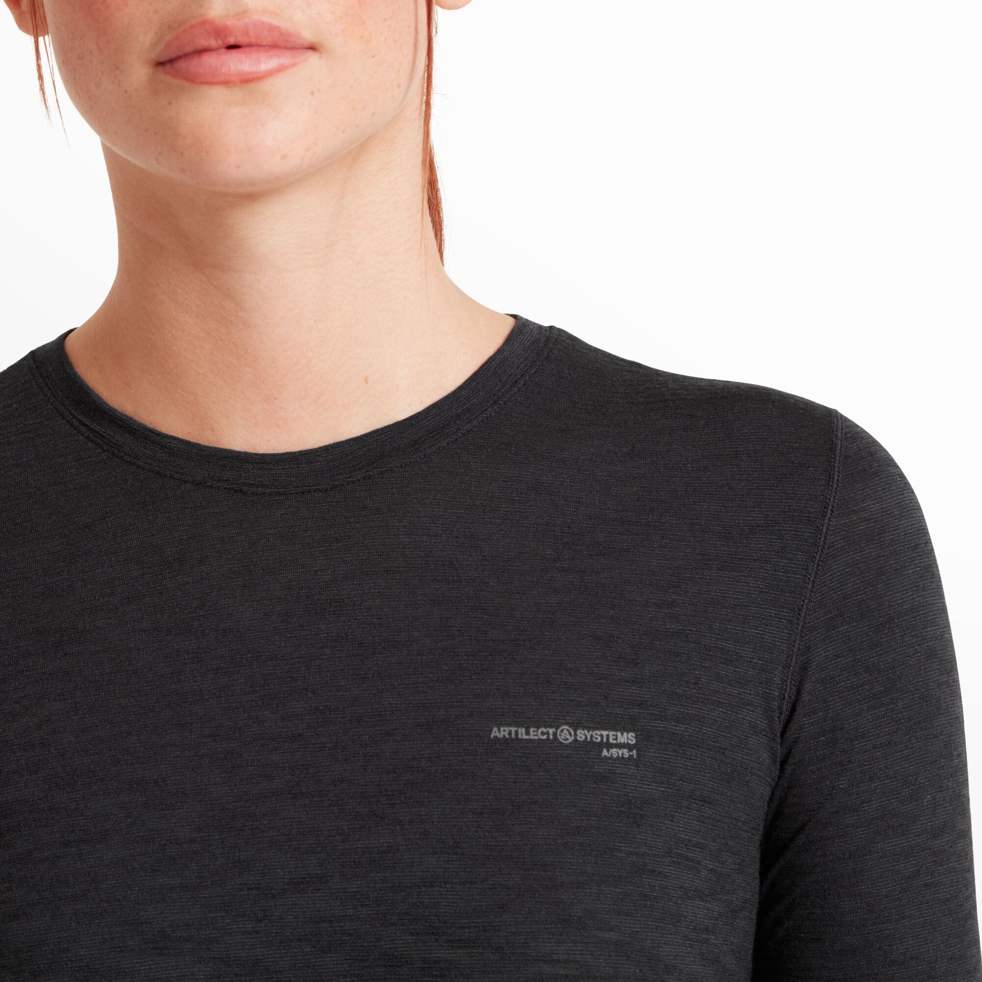 A close-up of the upper chest area of the Artilect Performance Womens Sprint Long Sleeve Tee Black, focusing on the neckline and logo. The fabric texture is slightly heathered, and the small "ARTILECT SYSTEMS A/SYS-1" logo is printed in a subtle gray on the left chest. The round neckline has fine stitching, giving it a clean finish.