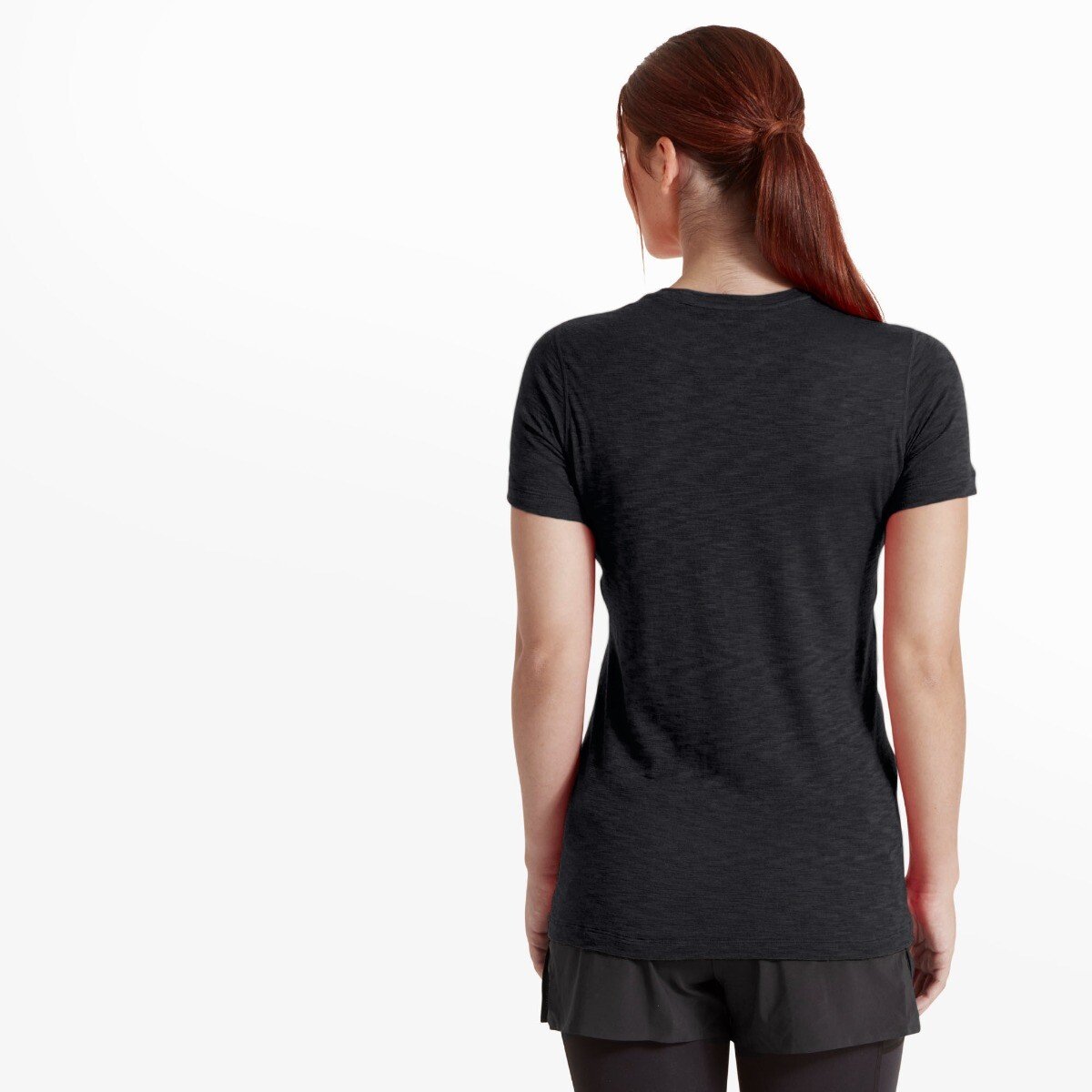 A rear view of the same model wearing the Artilect Performance Womens Sprint Tee Black, highlighting the elongated back hem and clean, minimal seam construction. The tee fits snugly through the back and shoulders, allowing a full view of the shirt’s form and fabric drape. Her hair is pulled back into a ponytail.