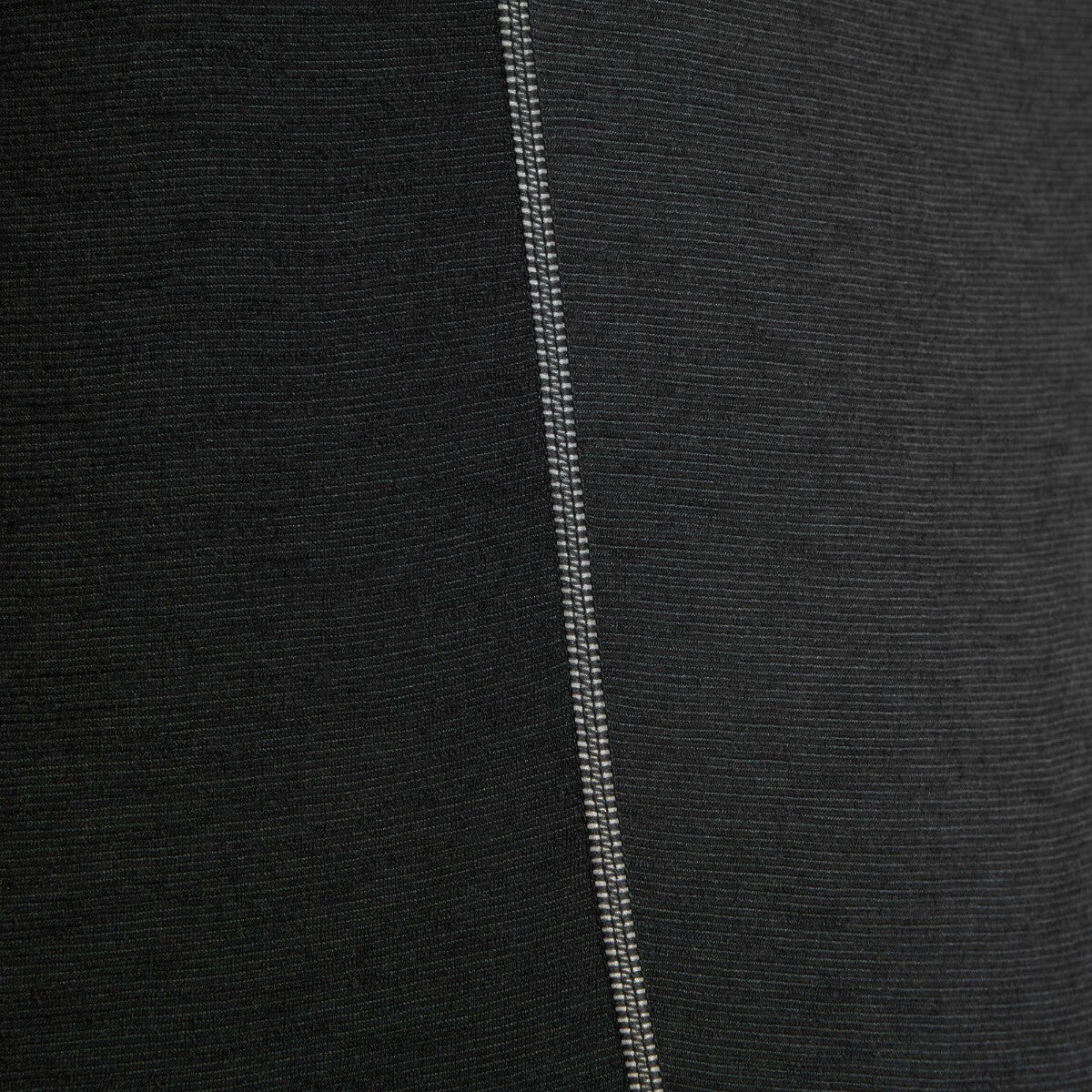 An extreme close-up of the Artilect Performance Womens Sprint Tee Black's side seam, showing the intricate flatlock stitching detail in contrasting white thread against the black fabric. The material has a slightly ribbed texture, showcasing the technical quality and fine weave of the performance fabric.
