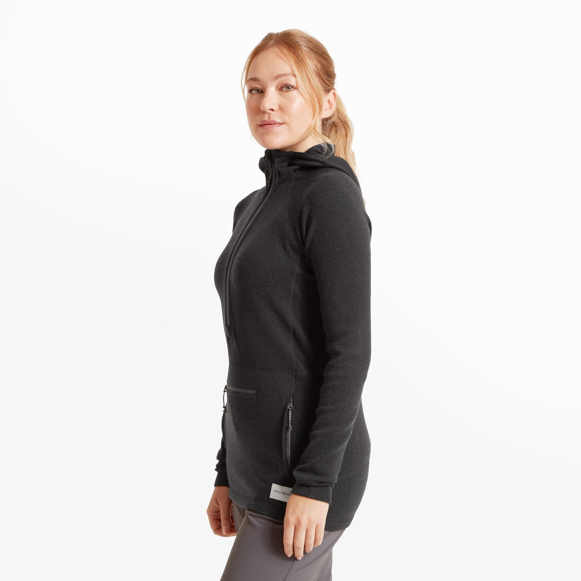 Side profile of the same woman modeling the Artilect Performance Womens Sundown 250 Half Zip Top Black. The length of the garment extends past the hips, and two front zippered pockets are visible. The hood lies flat against the back.